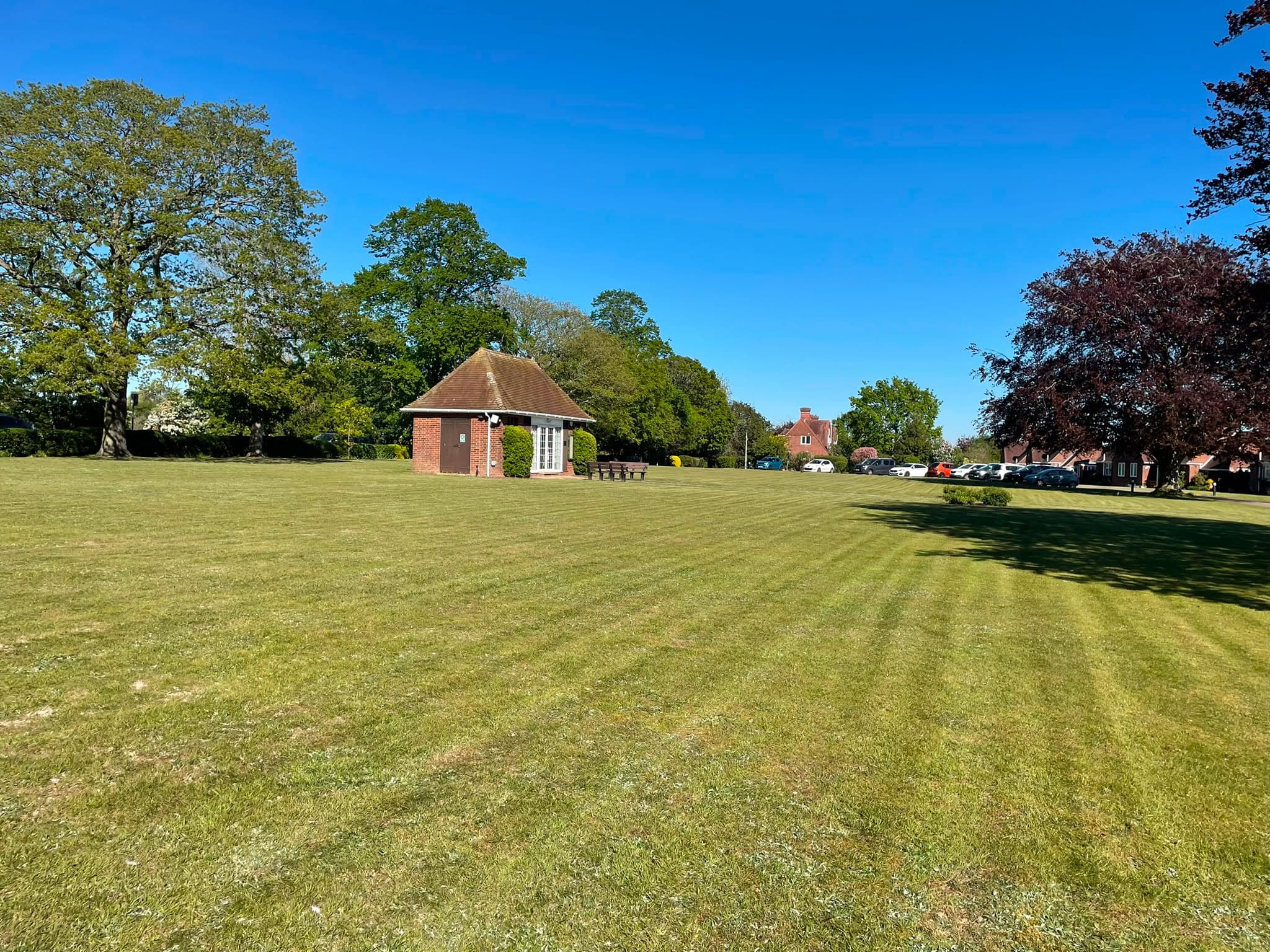 A large open green freshly mown into wide stripes, with a small brick pavilion and mature trees along the far edge